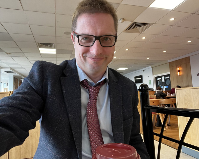 Photo of me sat in a claret and blue tweed, claret tie, blues shirt, and Burnley socks at knutsford services - nursing a cappuccino from Pret