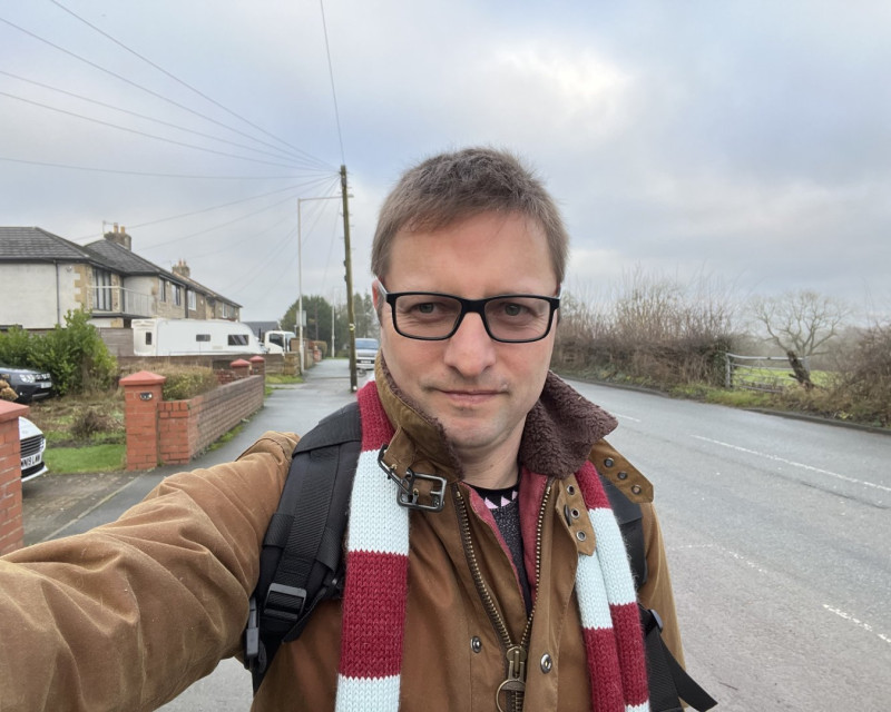 Me stood in Burnley wearing claret and blue scarf knitted by @craftedbypanda and Barbour Jacket.  Houses to the left, fields to the right with a road running through towards Pendle Hill under light grey sky.