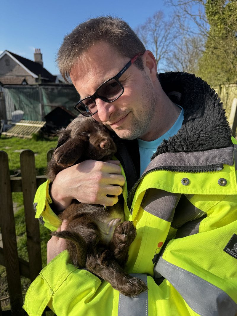 Picture of me, in hi-vis jacket, hugging our new cocker spaniel puppy, Ruaridh, on a sunny day, with neighbouring farmhouse beyond.