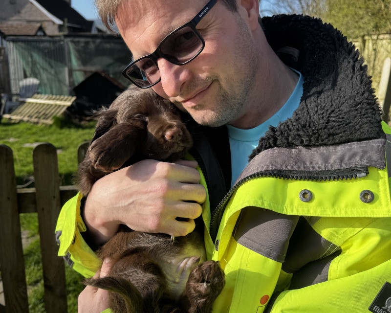 Picture of me, in hi-vis jacket, hugging our new cocker spaniel puppy, Ruaridh, on a sunny day, with neighbouring farmhouse beyond.