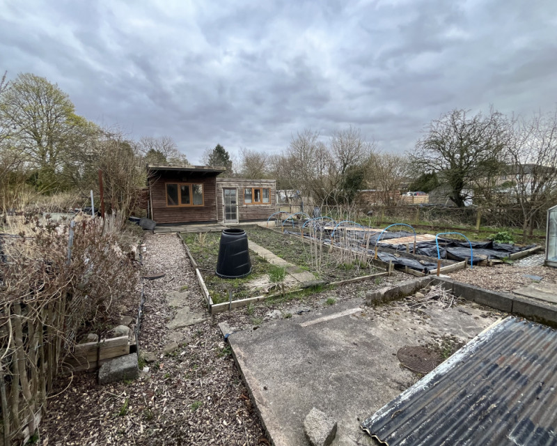 A picture looking across our allotment from one corner with a wide vision lens.  There are two sheds with a substantial verandah to the rear, a greenhouse to the right, and four substantial beds for growing across the plot.