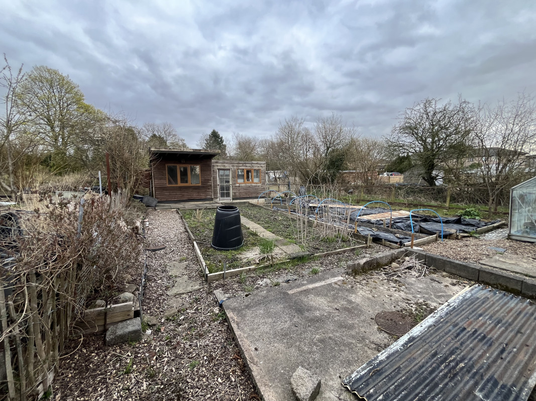A picture looking across our allotment from one corner with a wide vision lens.  There are two sheds with a substantial verandah to the rear, a greenhouse to the right, and four substantial beds for growing across the plot.