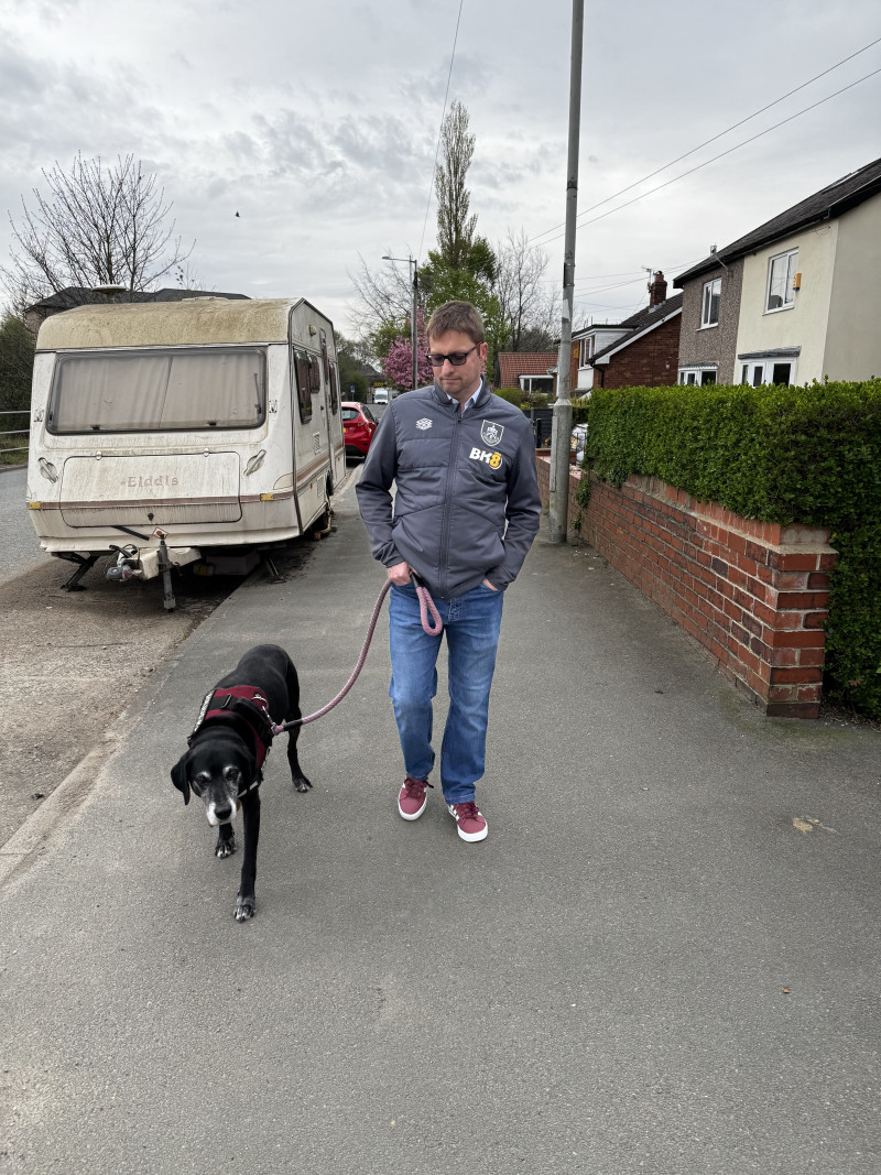 Old black pointer walks with me dressed in claret shoes, jeans, and a grey jacket, with a caravan covered in algae beyond