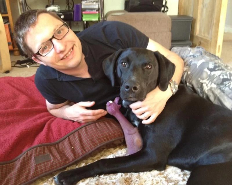 Picture of a black pointer in a group hug on the floor with his human, and his purple rubber dinosaur.